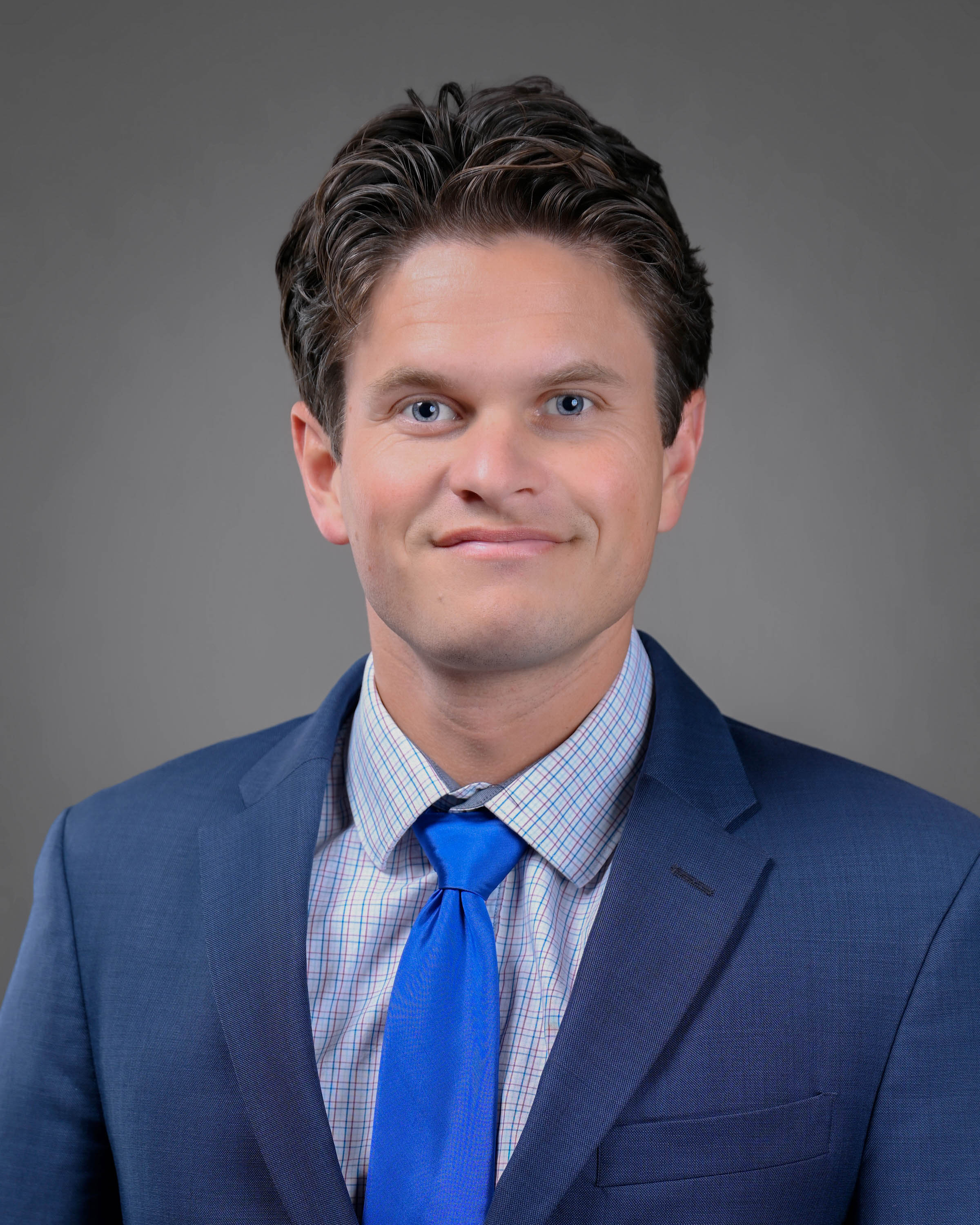 Clean shaven man with dark brown hair smiling in a blue suit with lighter blue tie